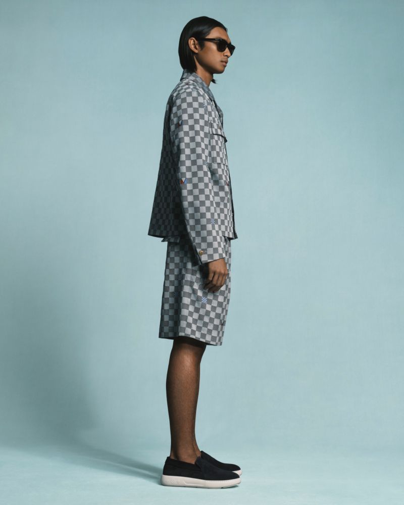 Studio shot of adult male fashion model in grey checkered jacket and shorts with black shoes, standing in profile against light blue background.