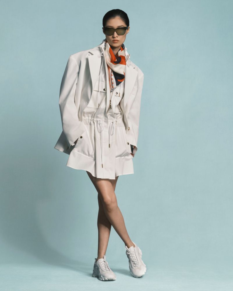 Woman in white jacket, top, skirt, sneakers and sunglasses in a full shot on a light blue background in a fashion studio shoot.