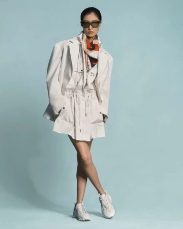 Woman in white jacket, top, skirt, sneakers and sunglasses in a full shot on a light blue background in a fashion studio shoot.