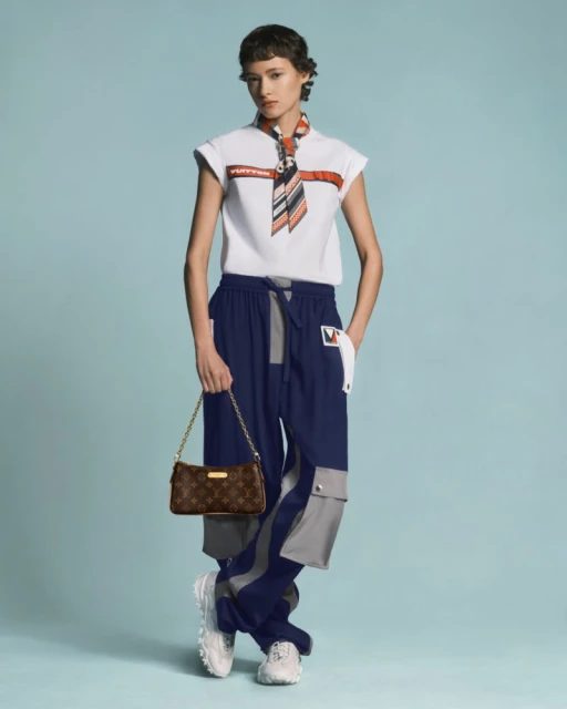 Fashion shot of a slender woman in a white tee, blue cargo pants, and white sneakers, carrying a brown monogrammed purse on a blue backdrop.