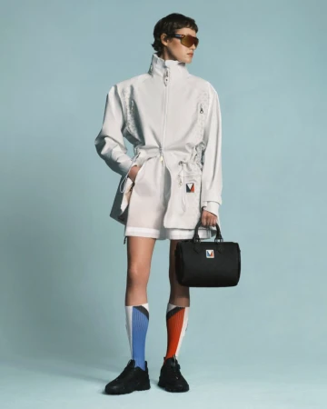 Fashion shot featuring a person in a white track suit and black shoes holding a black bag against a light blue background.