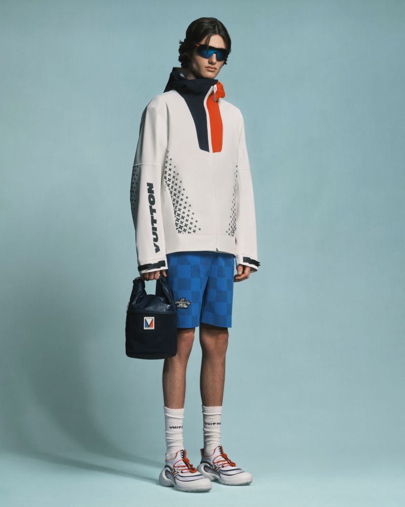 A full shot of a male model wearing a white jacket with blue shorts and white sneakers, against a light blue background.
