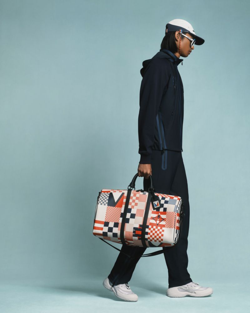 A male model walks against a light blue backdrop, carrying a patterned duffel bag and wearing a matching navy hooded tracksuit and cap.