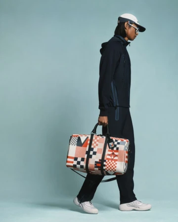 A male model walks against a light blue backdrop, carrying a patterned duffel bag and wearing a matching navy hooded tracksuit and cap.