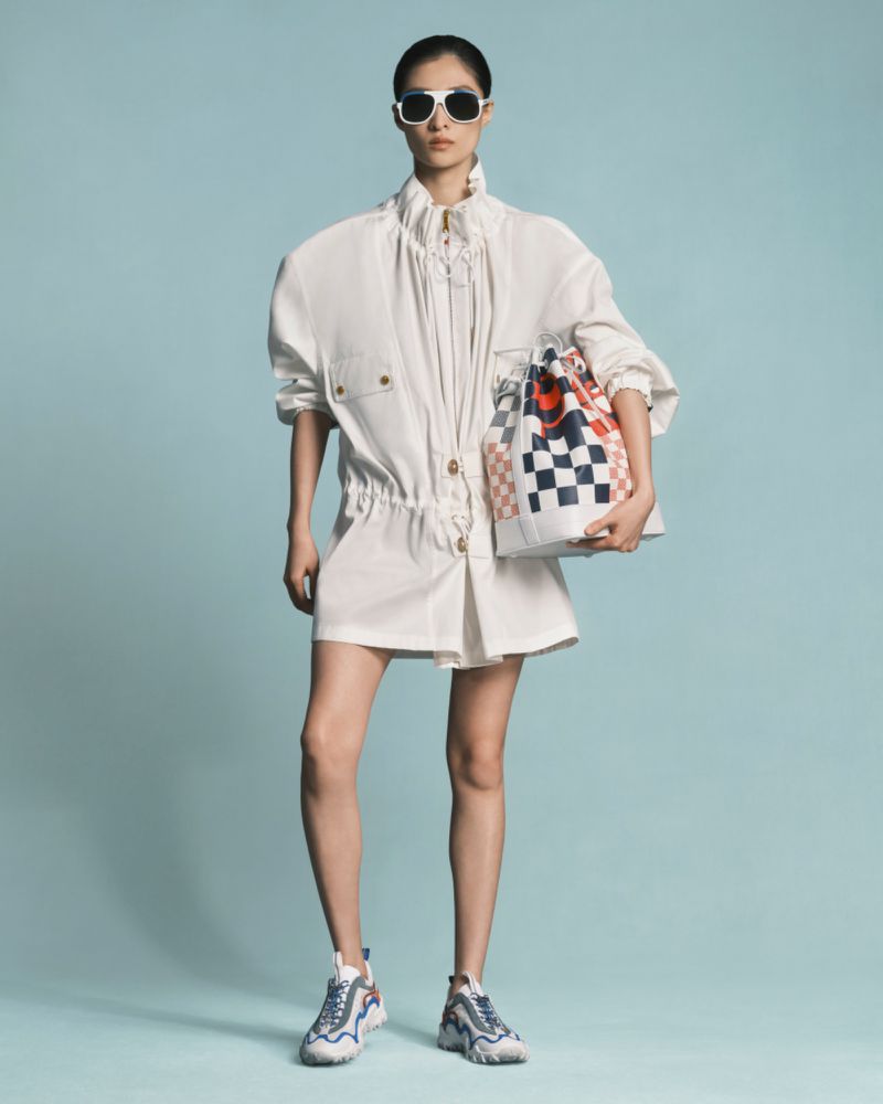 Fashion shot of an Asian woman wearing an all-white outfit and sunglasses holding a checkered bucket bag on a blue background.