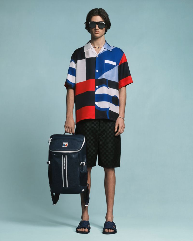 Full-shot studio image of a male model in patterned shirt and shorts holding a blue backpack, wearing sunglasses and sandals.