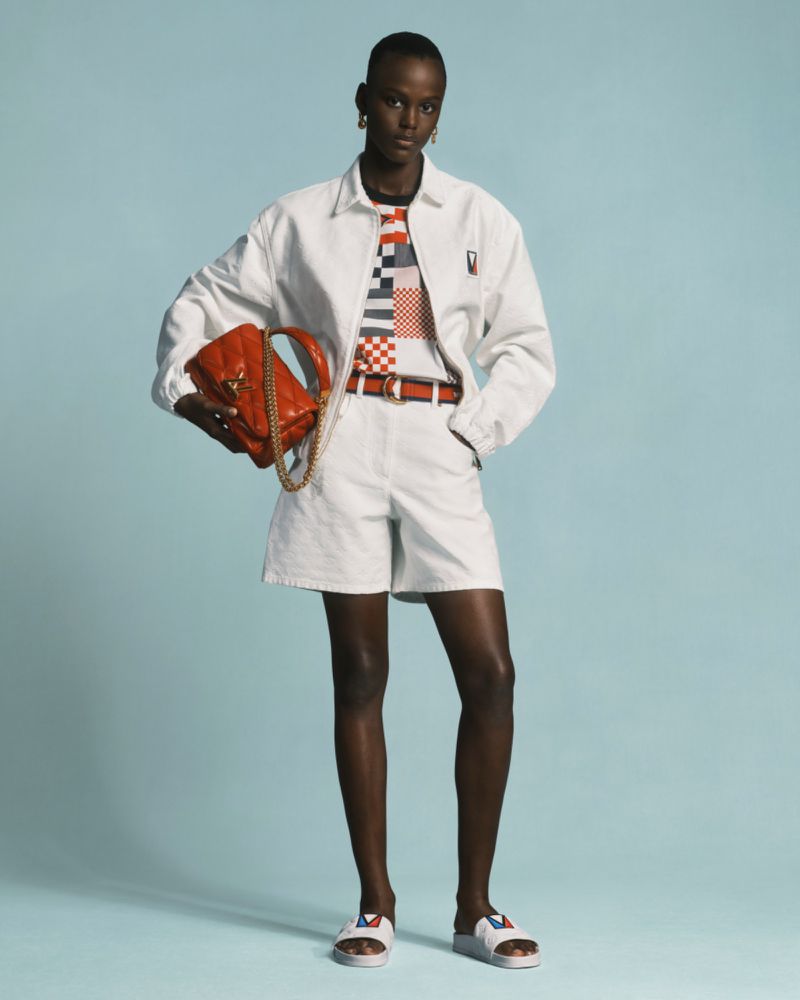 Woman in white jacket and shorts poses with orange purse against a light blue backdrop in a fashion editorial studio shot.
