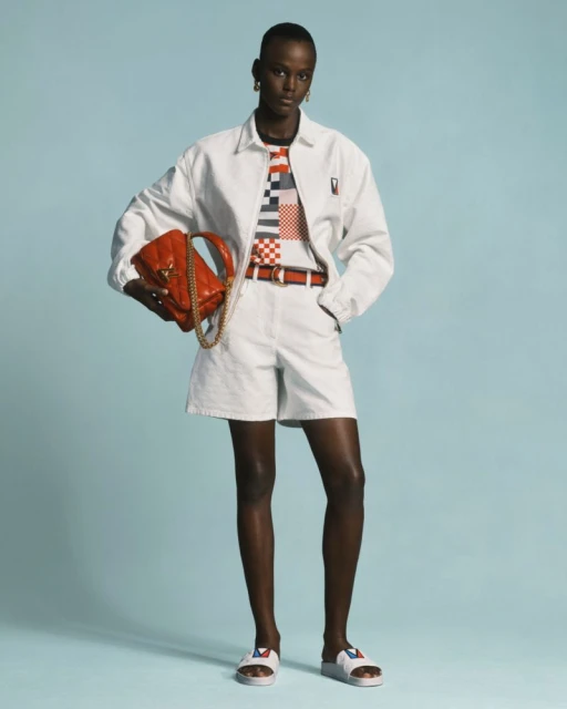 Woman in white jacket and shorts poses with orange purse against a light blue backdrop in a fashion editorial studio shot.