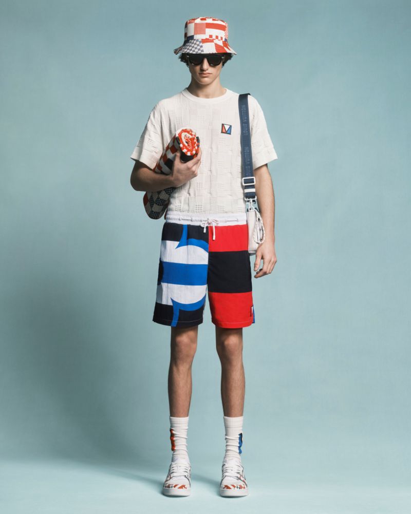 Male fashion model in a bucket hat, patterned shorts, and t-shirt, carrying a striped bag, posing against a light blue backdrop in a studio.
