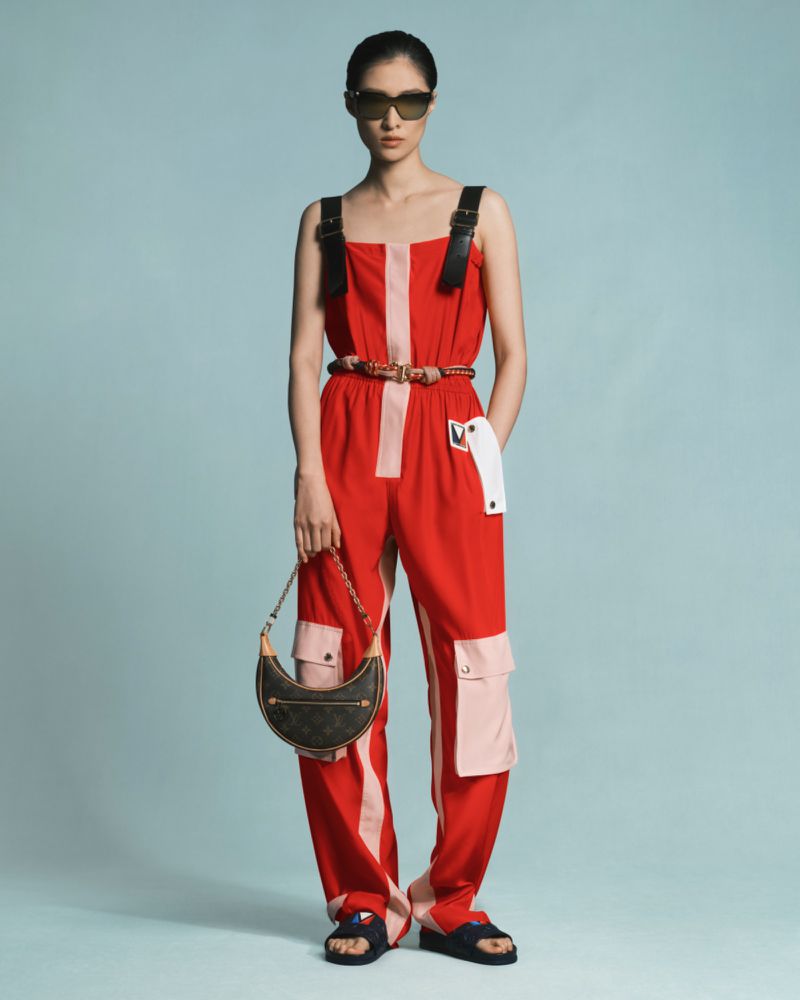 A woman models red color-blocked jumpsuit with black straps and sunglasses in a studio with a light blue backdrop, holding a brown purse.
