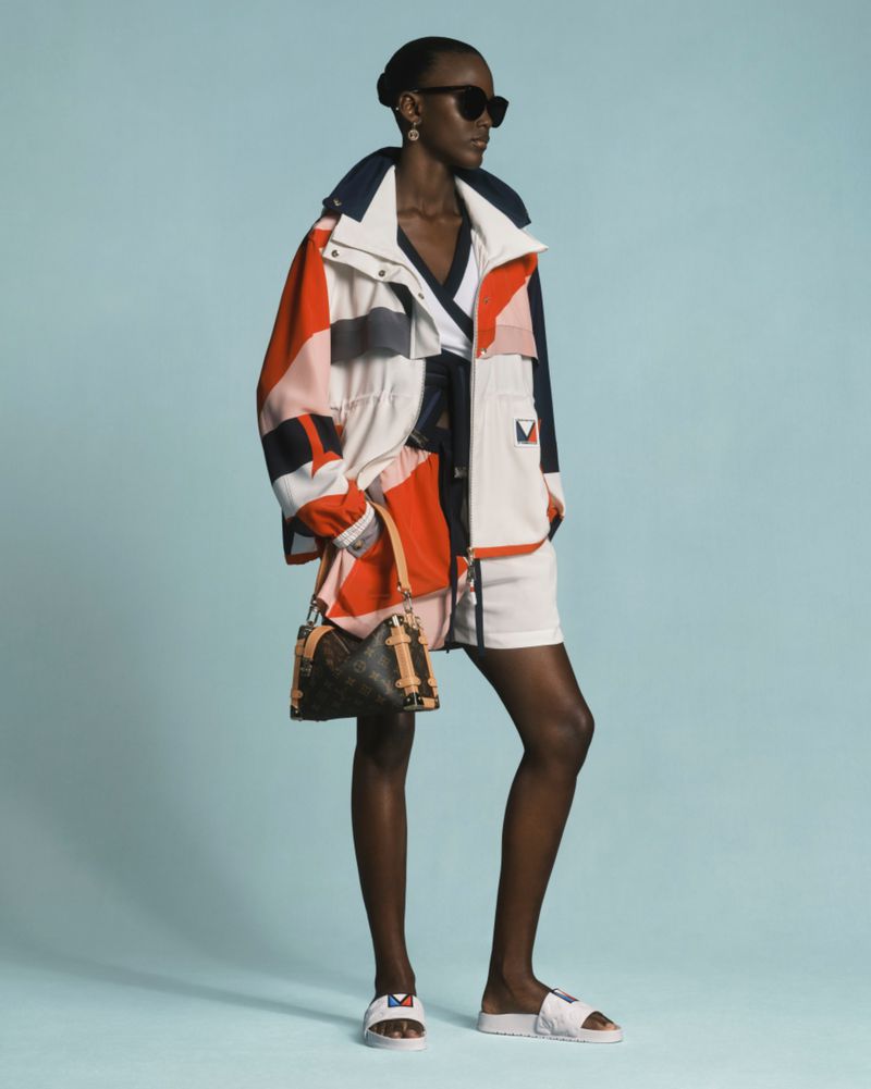 Fashionable black female model in sunglasses, jacket, and shorts, holding handbag in a brightly lit studio against a blue backdrop.