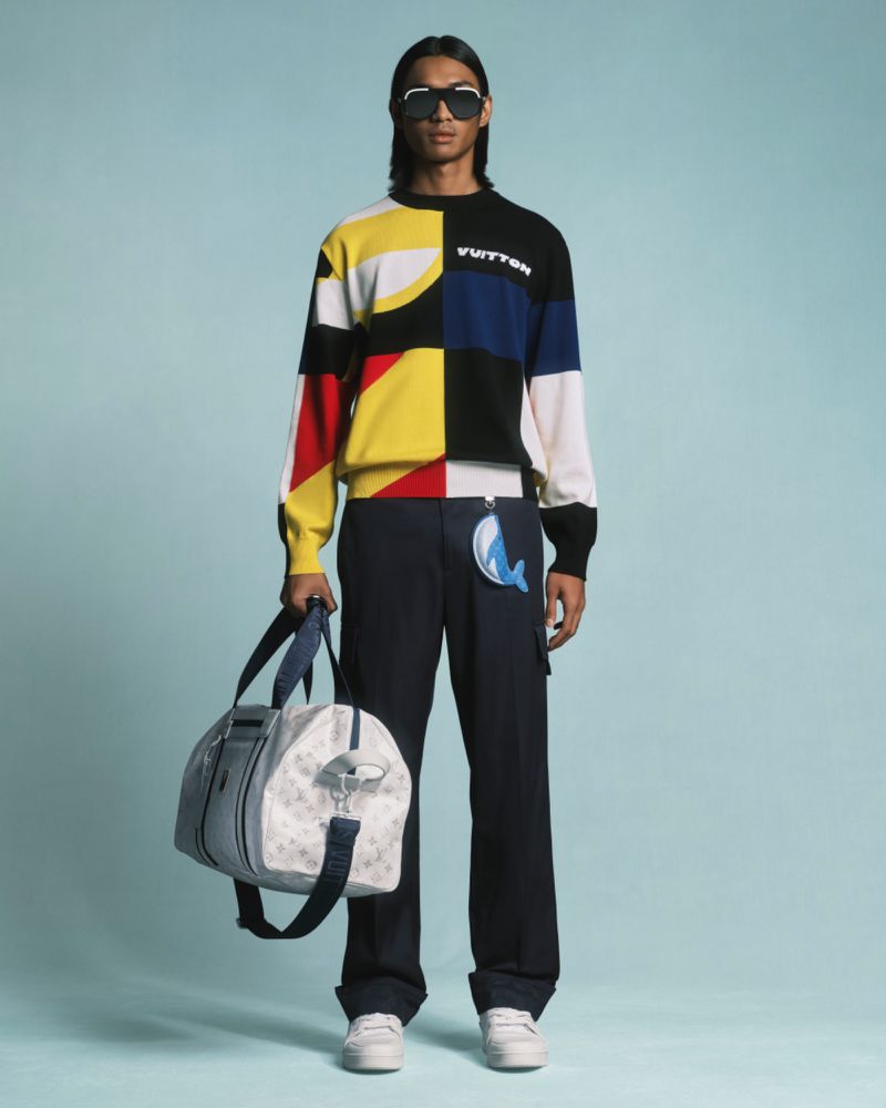 Male model in colorful patchwork sweater and wide-leg pants, holding patterned duffel bag in a studio setting with blue backdrop.