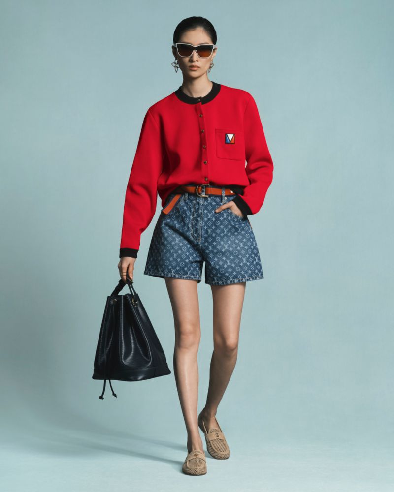 An Asian fashion model poses in a red cardigan, denim shorts with a logo pattern, and loafers, carrying a black drawstring bag.