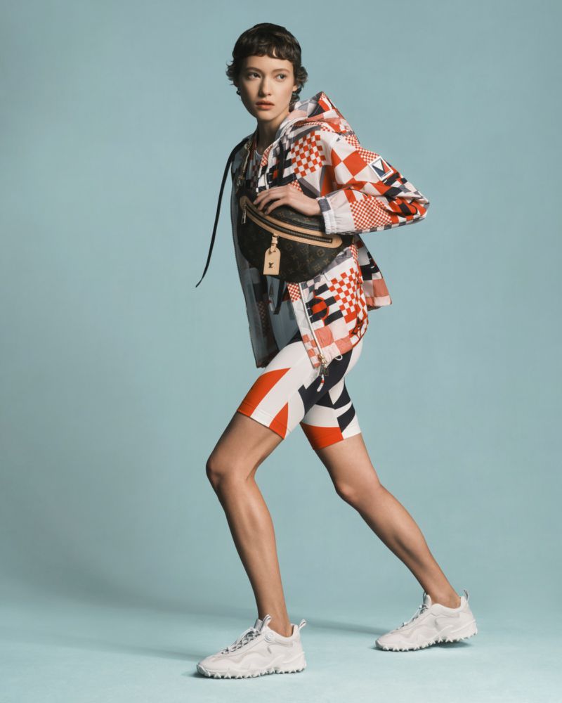 A woman models a patterned jacket and shorts, accessorized with a belt bag and white sneakers, set against a serene light blue background.