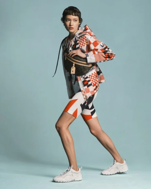A woman models a patterned jacket and shorts, accessorized with a belt bag and white sneakers, set against a serene light blue background.