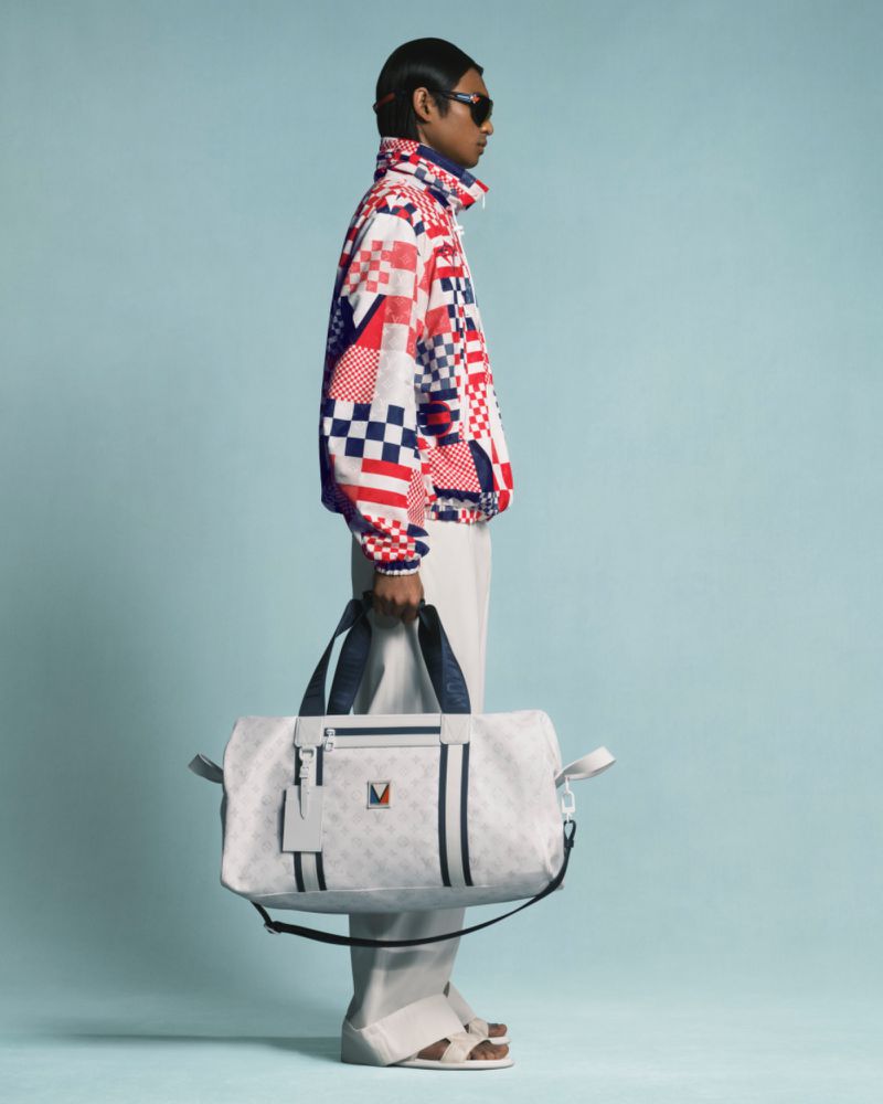 Stylish man in patterned jacket holds white duffel bag against light blue background in a full-length fashion shot.