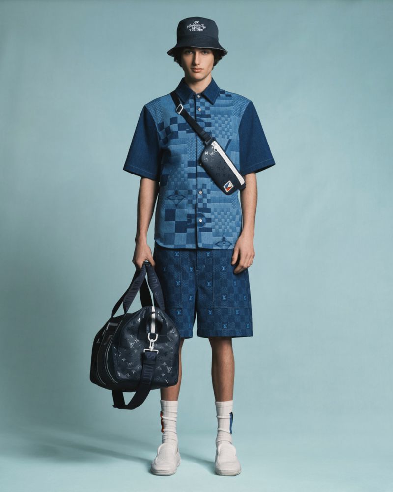 Man in blue patterned shirt, shorts and bucket hat, holding duffel bag in a studio shot with a light blue background.