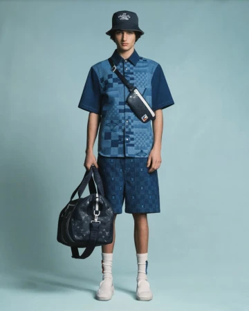 Man in blue patterned shirt, shorts and bucket hat, holding duffel bag in a studio shot with a light blue background.