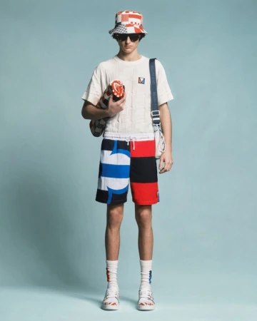 Male model in a patterned bucket hat, white knit shirt, colorblock graphic shorts, and sunglasses against a light blue studio backdrop.