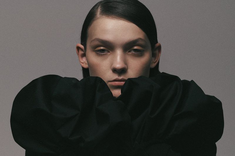 Close-up fashion portrait of a young adult woman with sleek, dark hair, wearing a voluminous black top against a muted grey backdrop.
