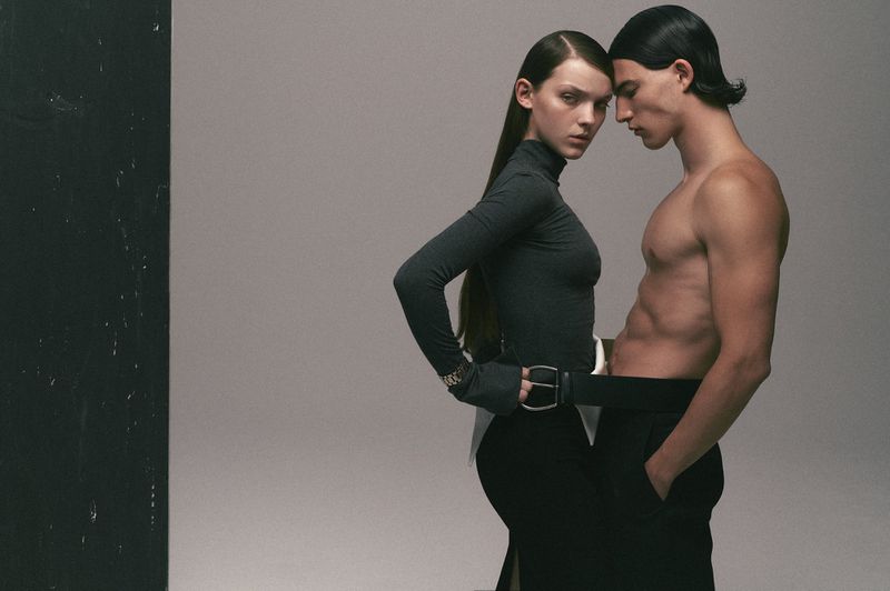 Fashion shot featuring a shirtless male model with abs and a female model in studio, both wearing black pants and posing.