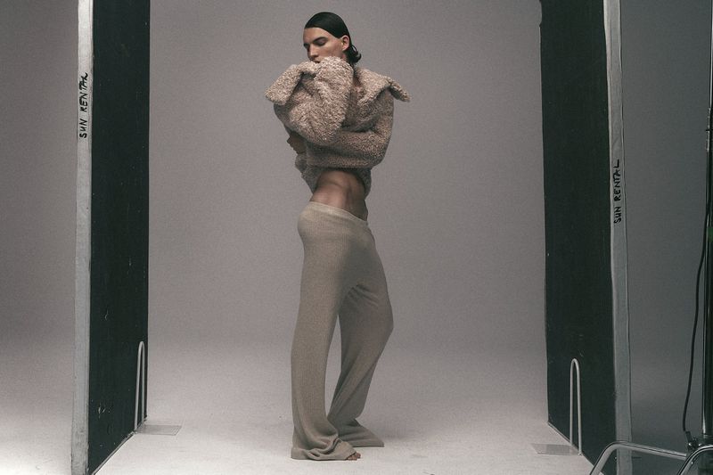 Fashion portrait of a slender adult man in a furry top and trousers, posing in a photo studio with 'SUN RENTAL' visible on equipment.