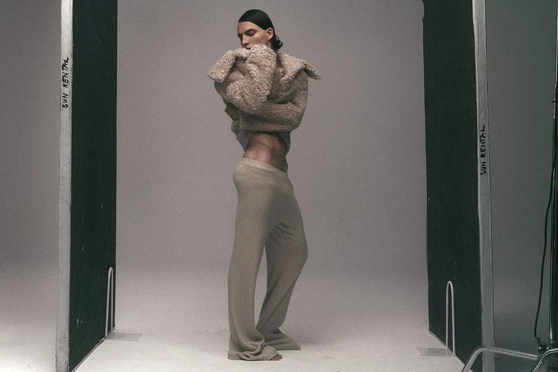 Fashion portrait of a slender adult man in a furry top and trousers, posing in a photo studio with 'SUN RENTAL' visible on equipment.