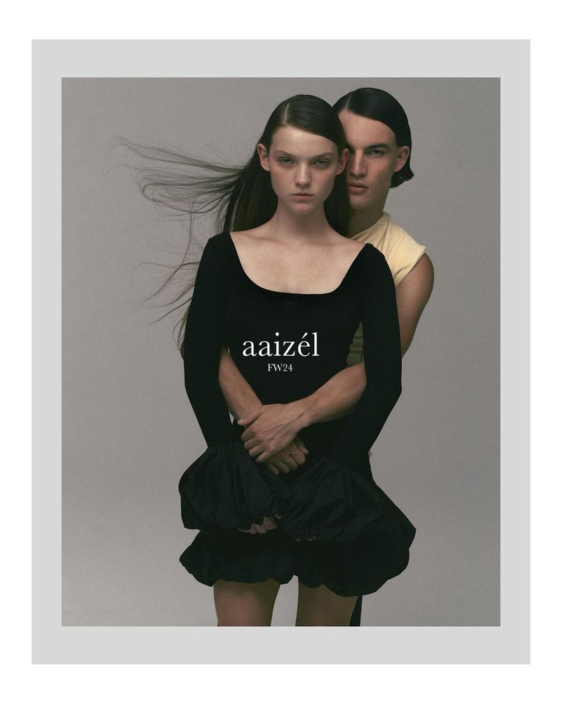A model couple poses in front of a gray background. The female model wears a black dress, while the male model is slightly behind her.