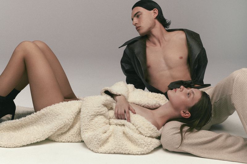 A fashion image features a young adult female lying down in an off-white coat and a young adult male sitting up in a black coat.