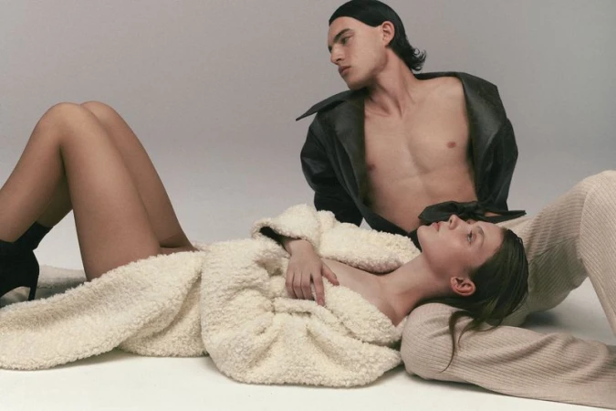 A fashion image features a young adult female lying down in an off-white coat and a young adult male sitting up in a black coat.