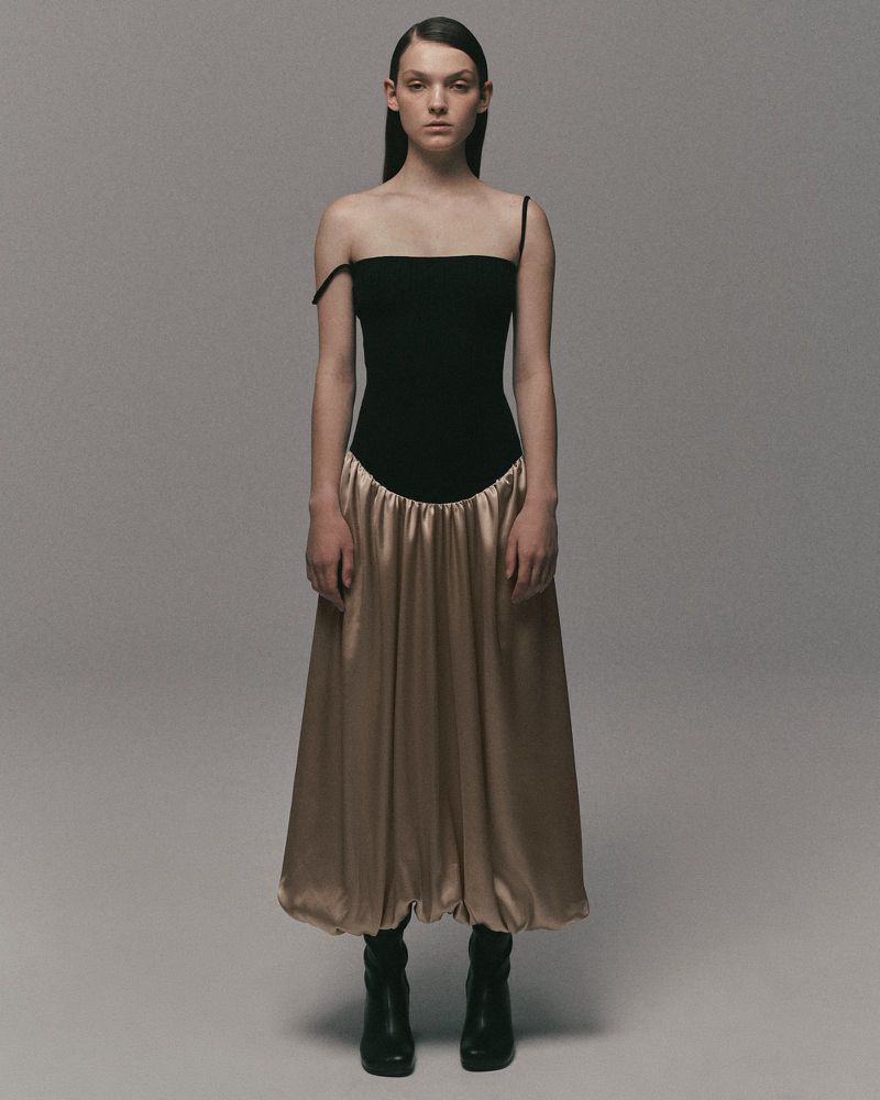 A full-length studio shot of a young woman in a black top, gold satin skirt, and black boots against a neutral backdrop, fashion portrait.