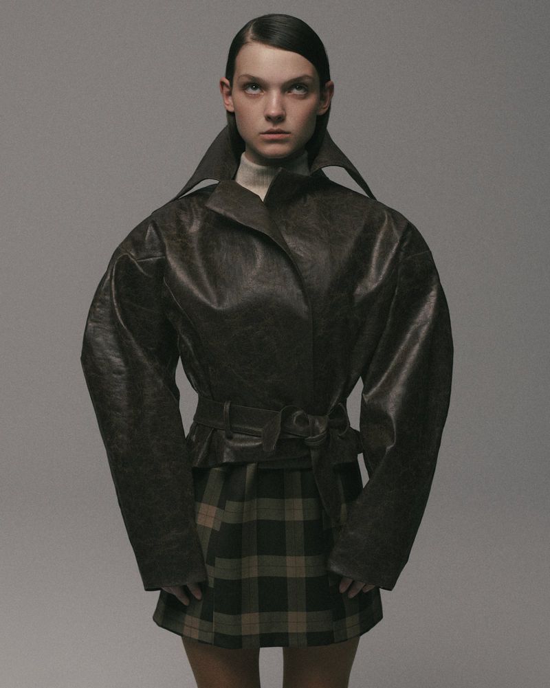 Fashion portrait of a young adult female model in a brown leather jacket and plaid skirt against a light gray studio backdrop.