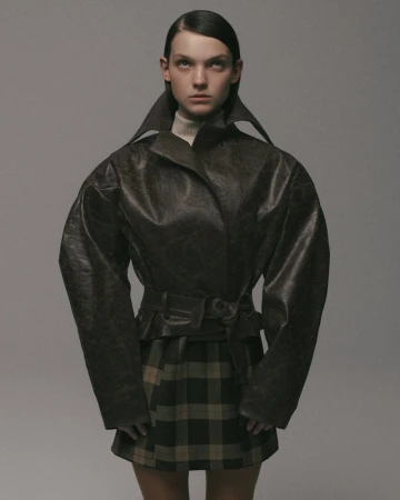 Fashion portrait of a young adult female model in a brown leather jacket and plaid skirt against a light gray studio backdrop.