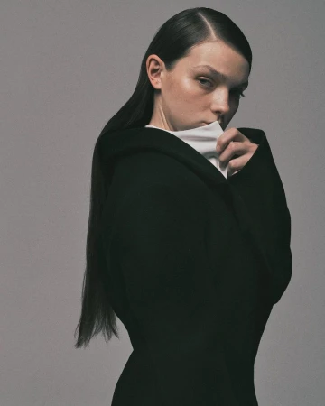 Model in a structured black coat with a high white collar looking over her shoulder against a grey studio backdrop.