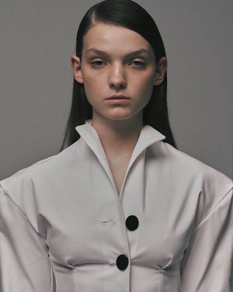 Headshot of a young woman with dark hair in a white collared jacket against a grey background, fashionable and minimalist.