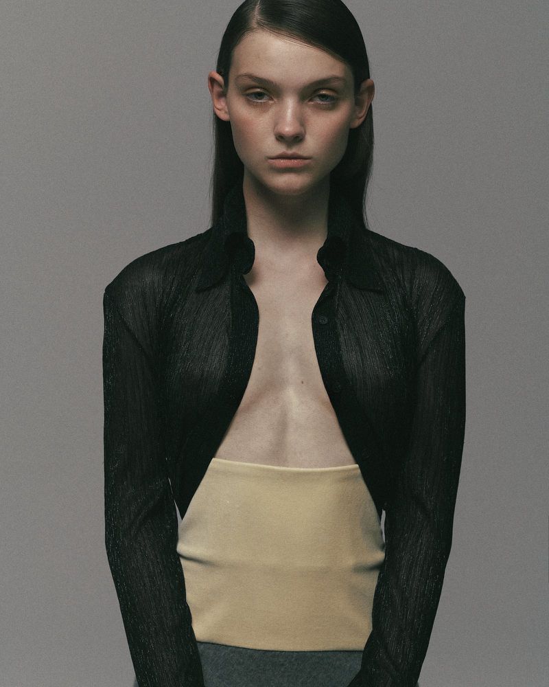 Attractive young woman poses in a studio wearing an open black cardigan, a tan top, and a grey skirt against a grey backdrop.