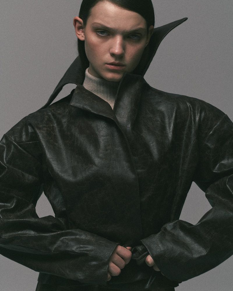 Fashion portrait of a woman wearing a black leather coat with a wide collar and a turtleneck, set against a gray studio background.