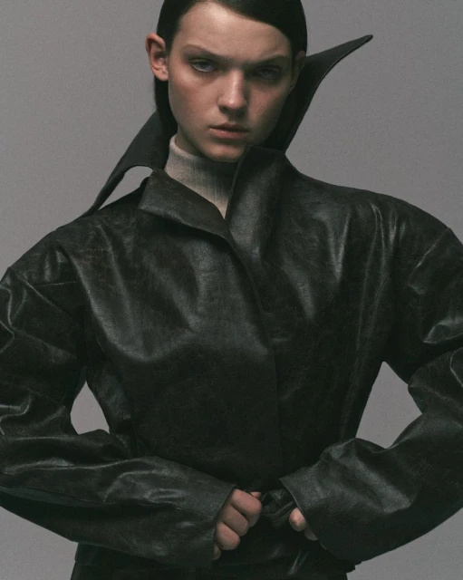 Fashion portrait of a woman wearing a black leather coat with a wide collar and a turtleneck, set against a gray studio background.