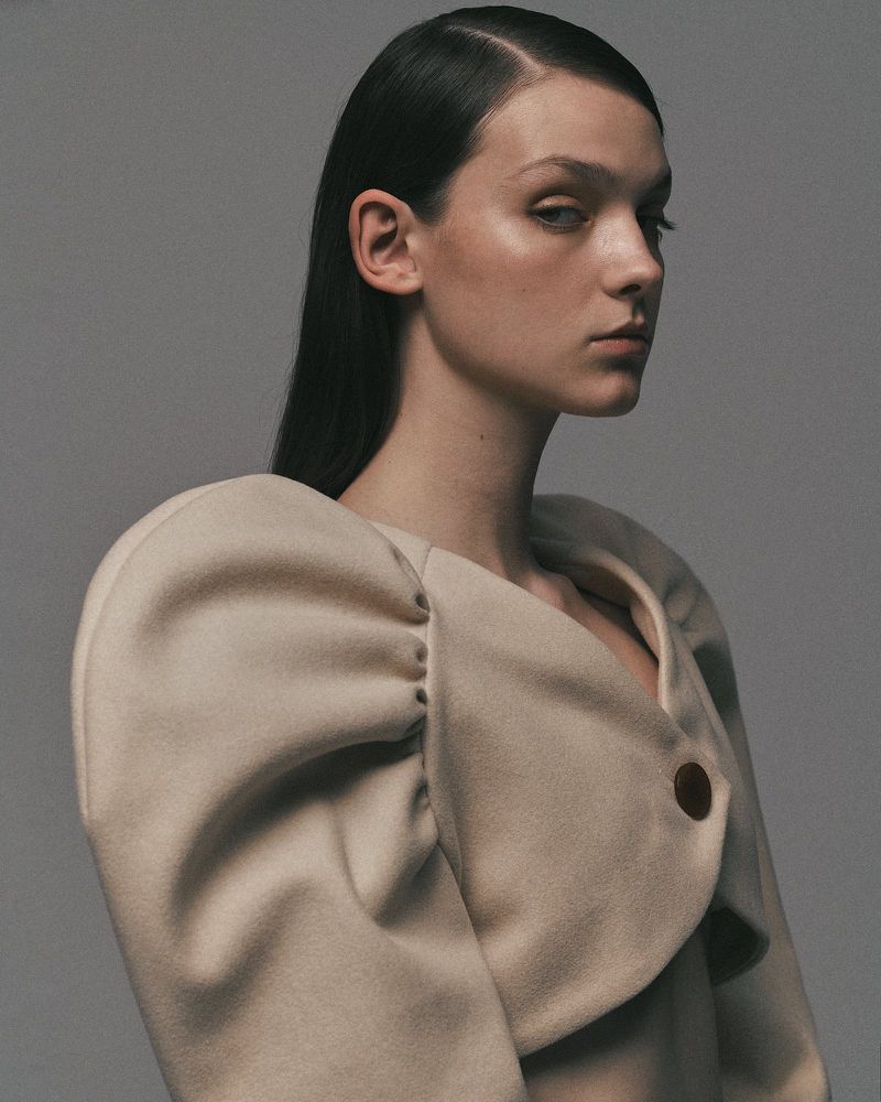 Woman with dark hair poses in a studio wearing a beige short coat with puffed sleeves, creating a fashion portrait.