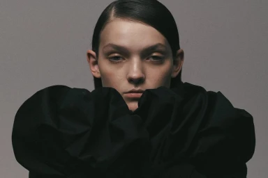 Close-up portrait of a woman with sleek dark hair wearing a voluminous black ruffled collar against a muted grey background.