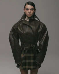 Model wearing a dark structured jacket with oversized shoulders and a belted waist over a plaid skirt against a plain grey background