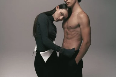A woman in a dark turtleneck resting her head on the shoulder of a shirtless man against a neutral grey studio background