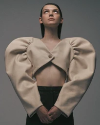 Model wearing a cropped beige structured top with exaggerated puffed shoulders against a grey seamless background.