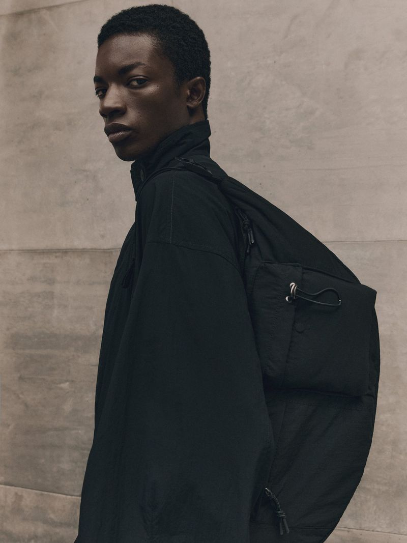 Studio shot of an African American male model in a black jacket and black bag, looking at the camera, against a neutral wall.