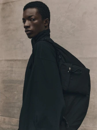 Studio shot of an African American male model in a black jacket and black bag, looking at the camera, against a neutral wall.