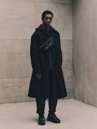 An African American man in black hooded sweatshirt, overcoat, pants, and sneakers poses against a neutral wall with a black bag.
