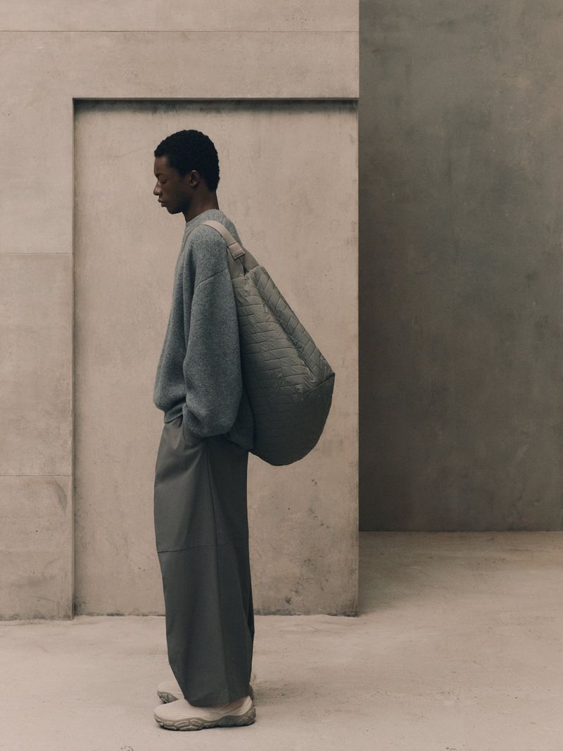 Fashion shot of a Black male model in a grey sweater and trousers carrying a quilted tote bag, posing against a concrete wall indoors.