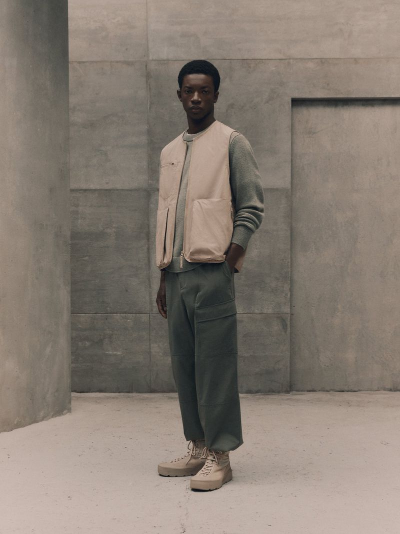 A lean man in an oyster-shell vest and green pants stands in a concrete space, emanating style and casual confidence in a neutral color palette.