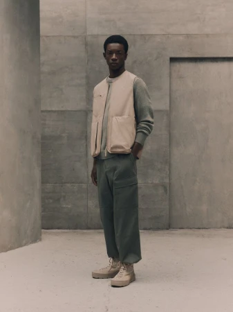 A lean man in an oyster-shell vest and green pants stands in a concrete space, emanating style and casual confidence in a neutral color palette.