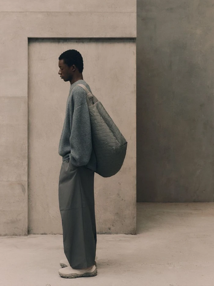 Model standing in profile wearing a grey sweater, wide trousers, and carrying a large shoulder bag against a neutral concrete wall.